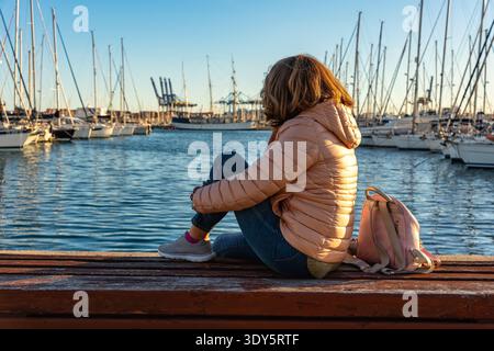 Frau, die bei Sonnenuntergang auf einer Holzbank ruht und auf die Boote im Hafen von Valencia blickt. Stockfoto