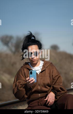 Junge Erwachsene Frau in lässiger Kleidung, die draußen ein Handy hält Stockfoto