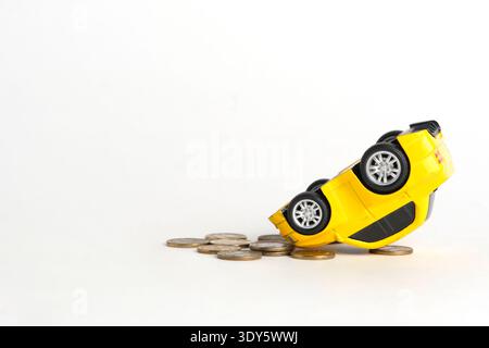 Nahaufnahme gelbes Spielzeugauto stürzte ab und lag auf der Seite auf kleinen Stapelmünzen, weißem Hintergrund Stockfoto