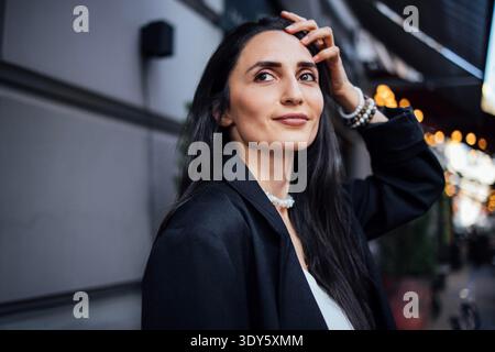 Stilvolle Frau mit langen dunklen Haaren, gekleidet in einem schwarzen Blazer, posiert selbstbewusst im Freien, umgeben von einer lebhaften urbanen Umgebung mit weichen Bokeh-Lichtern, die die Atmosphäre verbessern || Model erscheint Stockfoto