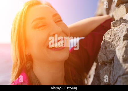Frau lacht Sonnenschein fröhliche reife Frau lächelt mit geschlossenen Augen, während sie sich während des Sonnenuntergangs an eine Steinmauer lehnt Stockfoto