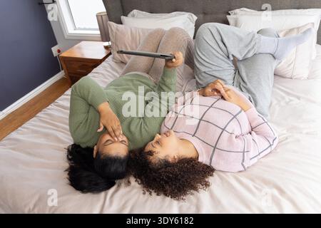 Zwei afroamerikanische Frauen, die im Schlafzimmer auf dem Bett liegen, Tablets halten und Pullover tragen. Kuschelig, paarweise, Gerät, Strickwaren, Lifestyle, Entspannung, Wärme || Model veröffentlicht Stockfoto