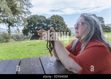 Seniorin findet Frieden und Spiritualität, meditiert mit gefesselten Händen und Gebetsperlen im Freien, nimmt Achtsamkeit und Entspannung in einer Ruhe auf Stockfoto