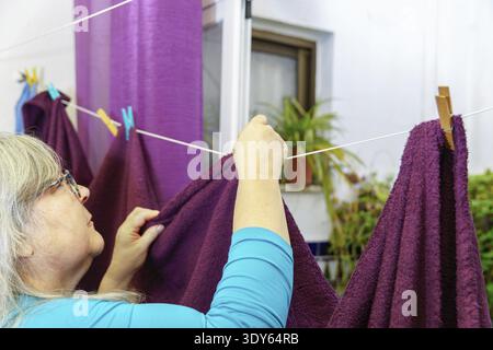 Reife Frau hängt frisch gewaschene lila Handtücher draußen an einer Wäscheleine mit Holz- und Plastik-Kleiderschränken, führt Hausarbeiten und Kuppel aus Stockfoto