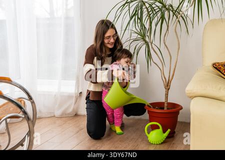 Frau und Kleinkind bewässern Zimmerpflanze mit Dose im Wohnzimmer. Stockfoto