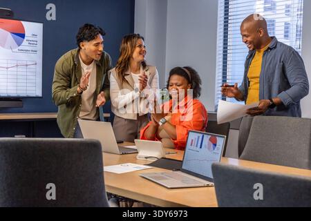 Verschiedene Kollegen applaudieren sitzende Frau in orangefarbener Bluse im Besprechungsraum mit Laptop-Tablet Stockfoto