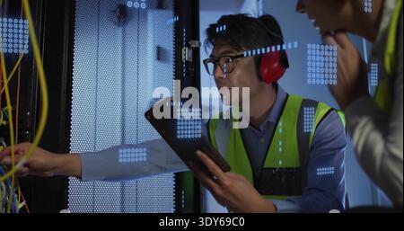 Inspektion asiatischer Technologie mit Tablet und Anlehnen in Server-Rack im Rechenzentrum, gelbe Kabel Stockfoto