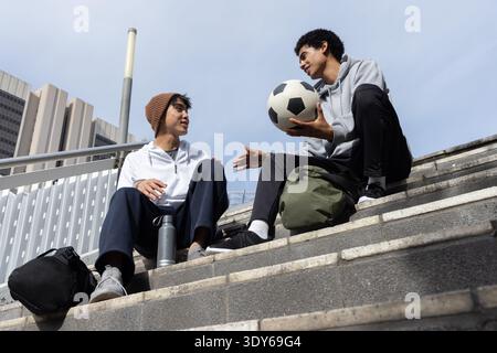 Verschiedene männliche Teenager sitzen auf Betontreppen und tragen Hoodies, die Fußball und Rucksäcke halten Stockfoto