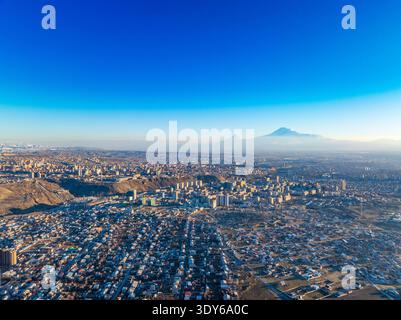 Luftaufnahme des weitläufigen Stadtbildes unter klarem blauem Himmel. Der ferne Berg Ararat erhebt sich über dem schneebedeckten Horizont. Die Stadt hat eine dichte Atmosphäre Stockfoto