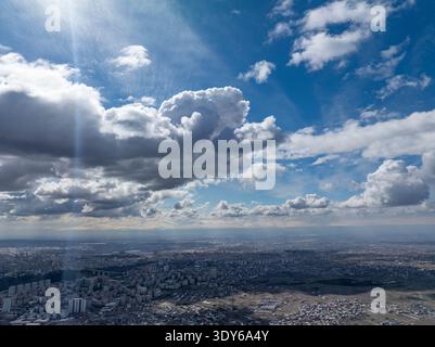 Aus der Vogelperspektive einer weitläufigen Stadt unter dramatischen Wolken. Sonnenlicht bricht durch und wirft Strahlen über die urbane Landschaft. Der Himmel dominiert die Szene mit flauschigem Whei Stockfoto