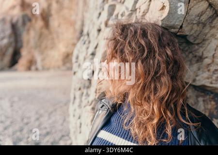 Lockige Frau, die sich an einem ruhigen, bewölkten Tag in einer Lederjacke an einer Steinmauer an einem Strand lehnt Stockfoto