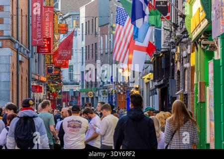 Gruppe von Menschen, die in einer geschäftigen Straße in der Nähe der Temple Bar in Dublin, Irland, spazieren gehen, mit Flaggen und Geschäften entlang der Straße Stockfoto