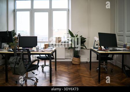 Leeres modernes Büro mit sauberen Schreibtischen mit Computermonitoren, organisiertem Arbeitsbereich und poliertem Holzboden, großen Fenstern für Tageslicht und Pflanzen im Innenbereich, die viel Grün verleihen Stockfoto