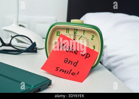 Eine rote Haftnotiz mit der Nachricht Wake up and Smile lehnt sich an einen grünen Wecker auf einem Nachttisch, in der Nähe befinden sich eine Brille und eine weiße Lampe Stockfoto