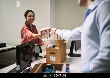 Junge Erwachsene kaukasische Frau lächelt, während sie jungen Erwachsenen kaukasischen Männern im modernen Büro Putzmittel überreicht, beide organisieren Gegenstände in Pappschachteln auf dem Schreibtisch Stockfoto