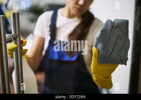 Junge Erwachsene kaukasische Frau, die die Glastür des Büros mit Mikrofasertuch und Sprühflasche reinigt, gelbe Gummihandschuhe trägt und sich darauf konzentriert, Flecken von der transparenten Oberfläche zu entfernen Stockfoto