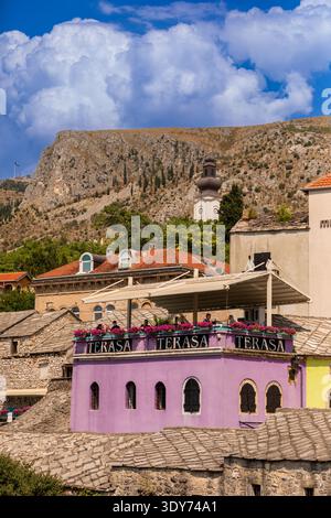 Das lebhafte, violette Dachcafé steht hoch zwischen Steinhäusern mit einem Kirchturm im Hintergrund in der Altstadt von Mostar Stockfoto