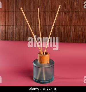 Eine minimalistische Komposition mit einem Glasdiffusor mit hölzernen Schilfen, vor einem strukturierten Bambusschirm und einem leuchtend rosa Hintergrund Stockfoto