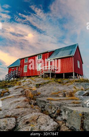 Rote Hütten sitzen auf felsigem Boden unter Wolken während der goldenen Stunde in Henningsvaer in der Nähe des Meeres auf den Lofoten-Inseln, Norwegen Stockfoto