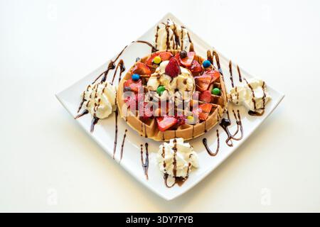 Köstliche Waffel mit Schokoladensoße und garniert mit Erdbeerscheiben, Eis, bunten Süßigkeiten und frischer Sahne Stockfoto