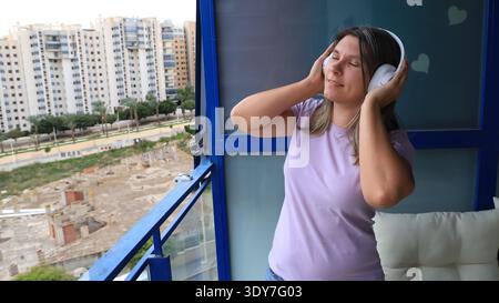 Frau mit Kopfhörern genießt Musik auf einem Balkon mit Blick auf städtische Gebäude Stockfoto