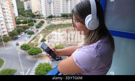 Frau mit Kopfhörern hört Musik auf dem Smartphone, während sie auf dem Balkon steht Stockfoto