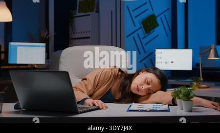 Geschäftsfrau schläft auf dem Schreibtisch, während sie spät in der Nacht in der Geschäftsstelle an den Rechnungslegungsstatistiken arbeitet. Überarbeiteter, gestresster erschöpfter Mitarbeiter am Arbeitsplatz, der Überstunden einnippt Stockfoto