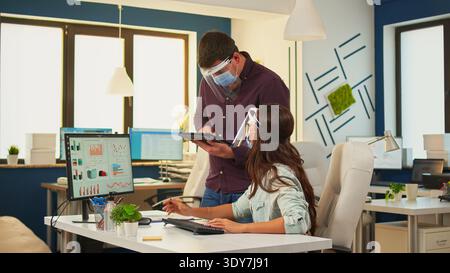 Geschäftsleute diskutieren Finanzstrategie und suchen Computer mit Schutzmaske in einem neuen normalen Bürozimmer. Das Multietnic-Team analysiert Diagramme, arbeitet zusammen und respektiert soziale Distanz. Stockfoto