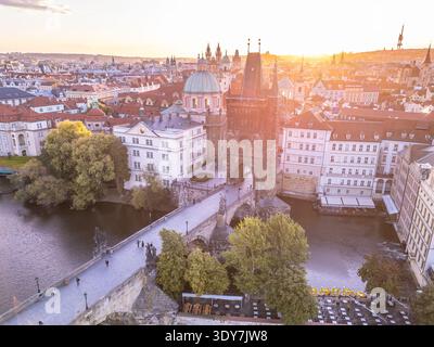 Dieser Blick aus der Vogelperspektive zeigt den Altstadtbrückenturm an der Karlsbrücke mit malerischen Gebäuden und den Fluss Moldau bei Sonnenuntergang. Man kann Leute sehen, die die Brücke entlang schlendern. Stockfoto