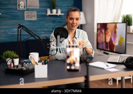 Bloggerin, die Buchrezension für ihren Online-Talk-Skow aufnimmt. Kreativer Vlogger, der ein Video-Blog-Konzept erstellt, spricht und betrachtet Smartphone auf einem Stativ im Heimstudio-Podcast mit moderner Ausrüstung. Stockfoto