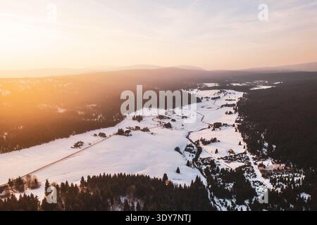 Aus der Vogelperspektive zeigt das Bergdorf Jizerka, das mit Schnee bedeckt ist. Die Landschaft ist weiß und grün, mit Bäumen und gewundenen Pfaden. Die Sonne untergeht über dem Dorf und den Bergen in der Nähe. Stockfoto