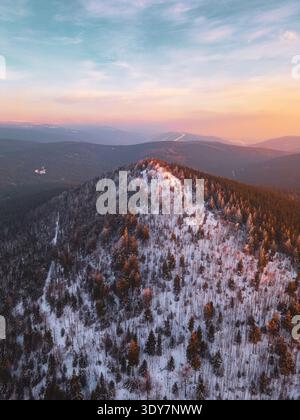 Aus der Vogelperspektive zeigt den Berg Bukovec im Isergebirge. Die Szene zeigt schneebedeckte Bäume und Hügel am späten Nachmittag. Die Sonne untergeht am Horizont und erzeugt warme Farben. Stockfoto