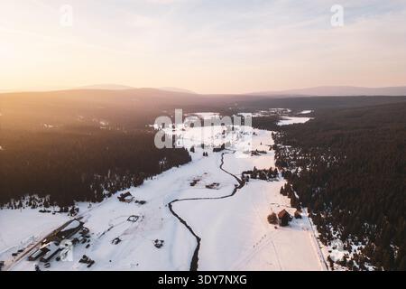 Aus der Vogelperspektive zeigt das Bergdorf Jizerka, das im Winter mit Schnee bedeckt ist. Bäume umgeben das Gebiet, während sich die Wege durch die weiße Landschaft schlängeln. Die Szene fängt die Schönheit der Saison ein. Stockfoto