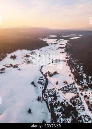 Das Bergdorf Jizerka zeigt eine schneebedeckte Landschaft von oben. Die Häuser sind von weißen Feldern und Bäumen umgeben. Das Sonnenlicht erzeugt im Winter sanfte Schatten über das Land. Stockfoto