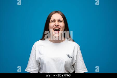 Junge Frau, die schreiend ist, Wut, Frustration und Stress ausdrückt, sich aufgeregt fühlt oder Schmerzen hat, vor blauem Studiohintergrund Stockfoto