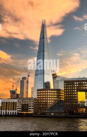 Die Szene zeigt den Shard, der über nahe gelegenen Gebäuden in London thront, während der Abendhimmel seine Farbe ändert. Kranmaschinen sind im Hintergrund zu sehen, was auf laufende Bauarbeiten hinweist. Stockfoto
