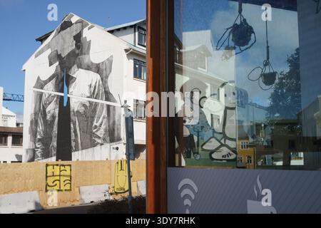 Großes einfarbiges Street Art Wandgemälde von INO, das neben einem farbenfrohen Charakterkunstwerk in einem Café-Fenster im Zentrum von Reykjavík, Island, reflektiert wird Stockfoto