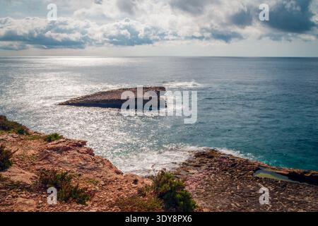 Sonnendurchflutete felsige Küste und kleine Insel unter bewölktem Himmel mit ruhigem blauem Meer und glitzerndem Horizont Stockfoto