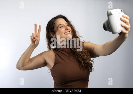 Lächelnde Frau, die einen glücklichen Moment mit sofortiger Kamera und Gesten des Friedenszeichens festhält Stockfoto