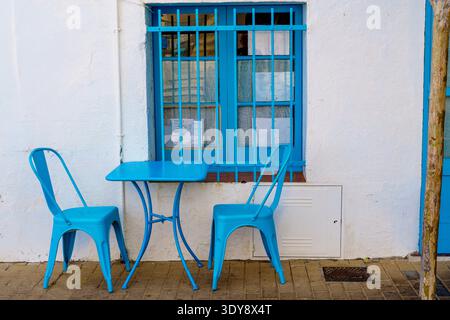 Zwei hellblaue Metallstühle und ein passender Tisch, der vor einer weißen Stuckwand mit einem blauen Fenster und vergitterten Scheiben steht, Sitzszene im Freien auf dem Bürgersteig Stockfoto