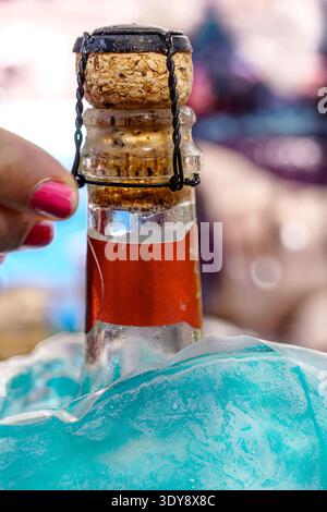 Nahaufnahme eines Sektflaschenkorkens mit Drahtkäfig, Hand mit rosa nagellack, der den Käfig hält, Flaschenhals in rote Folie gewickelt, Flasche gekühlt in bl Stockfoto