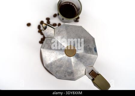 Blick von oben auf eine silberne Mokakanne auf weißem Hintergrund mit verstreuten Kaffeebohnen und einer Tasse gebrühten Kaffee, helle minimale Zusammensetzung Stockfoto