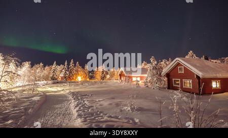Erleben Sie die bezaubernde Schönheit von Akaslompolo in Lappland, Finnland, wo leuchtende Nordlichter bezaubernde Holzhütten vor einer ruhigen Schneelandschaft bei Nacht erleuchten. Stockfoto