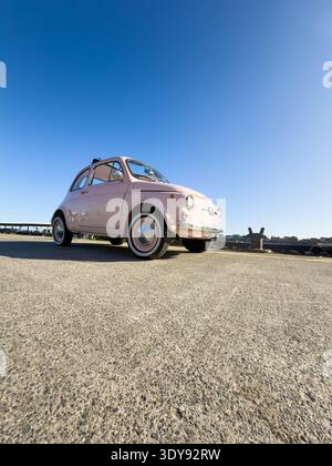 Niedrige Weitwinkelaufnahme eines klassischen Vintage-Fiat 500 aus den 1960er Jahren in hellrosa, geparkt auf Asphaltboden nahe der Hafenkante in North Shields Fish Quay, UK Stockfoto