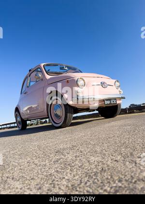 Niedrige Weitwinkelaufnahme eines klassischen Vintage-Fiat 500 aus den 1960er Jahren in hellrosa, geparkt auf Asphaltboden nahe der Hafenkante in North Shields Fish Quay, UK Stockfoto