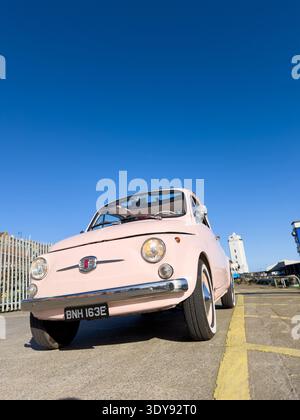 Niedrige Weitwinkelaufnahme eines klassischen Vintage-Fiat 500 aus den 1960er Jahren in hellrosa, geparkt auf Asphaltboden nahe der Hafenkante in North Shields Fish Quay, UK Stockfoto