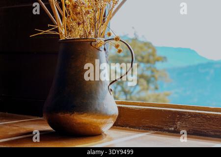 Metallvase mit getrockneten Blumen am Fenster bei Sonnenaufgang. Dekoration eines Hauses auf dem Land Stockfoto