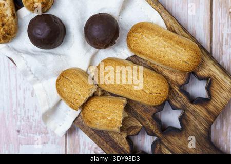 Verschiedene Kekse auf rustikalem Holzbrett: Marienkreuzfinger, kaputte Keksstücke und Kugeln mit Schokolade auf weißem Tuch über hellem Holztisch, Blick von oben. Stockfoto