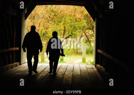 Ein erwachsenes Paar spaziert an einem Herbsttag durch eine überdachte Brücke, während die helle Szene sie in Sheffield, Massachusetts, in Silhouette wiedergibt Stockfoto