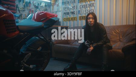 Motorradfahrerin plaudert mit der Biker Group auf dem Smartphone in der professionellen Garage. Junge Frau sitzt in der Nähe von Sportbikes und diskutiert auf Mobilgeräten die Details der zukünftigen Reise. Biker-Subkultur. Hochformat. Stockfoto